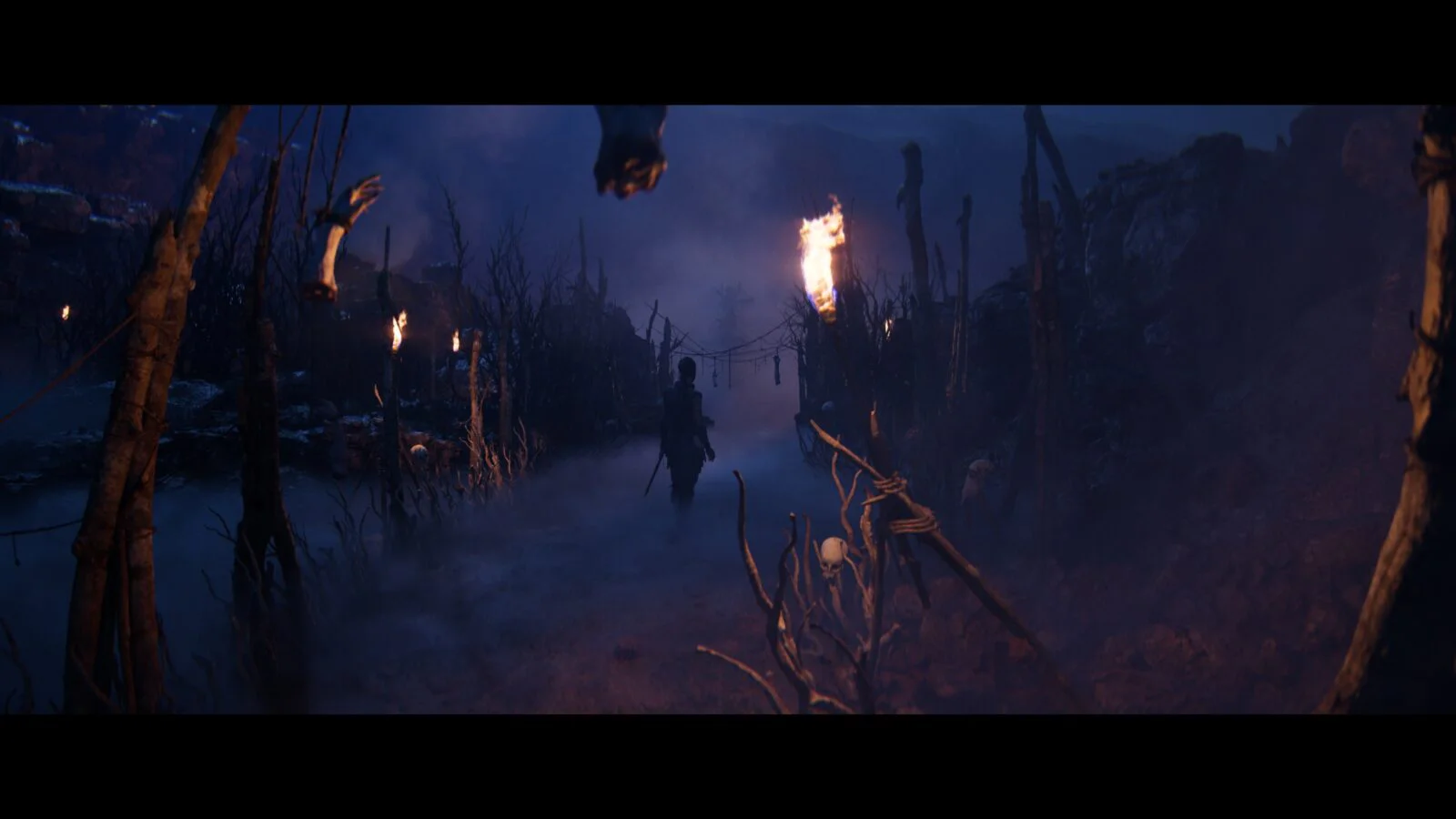 Senua идёт через мрачный туманный проход с факелами и костями в Hellblade II